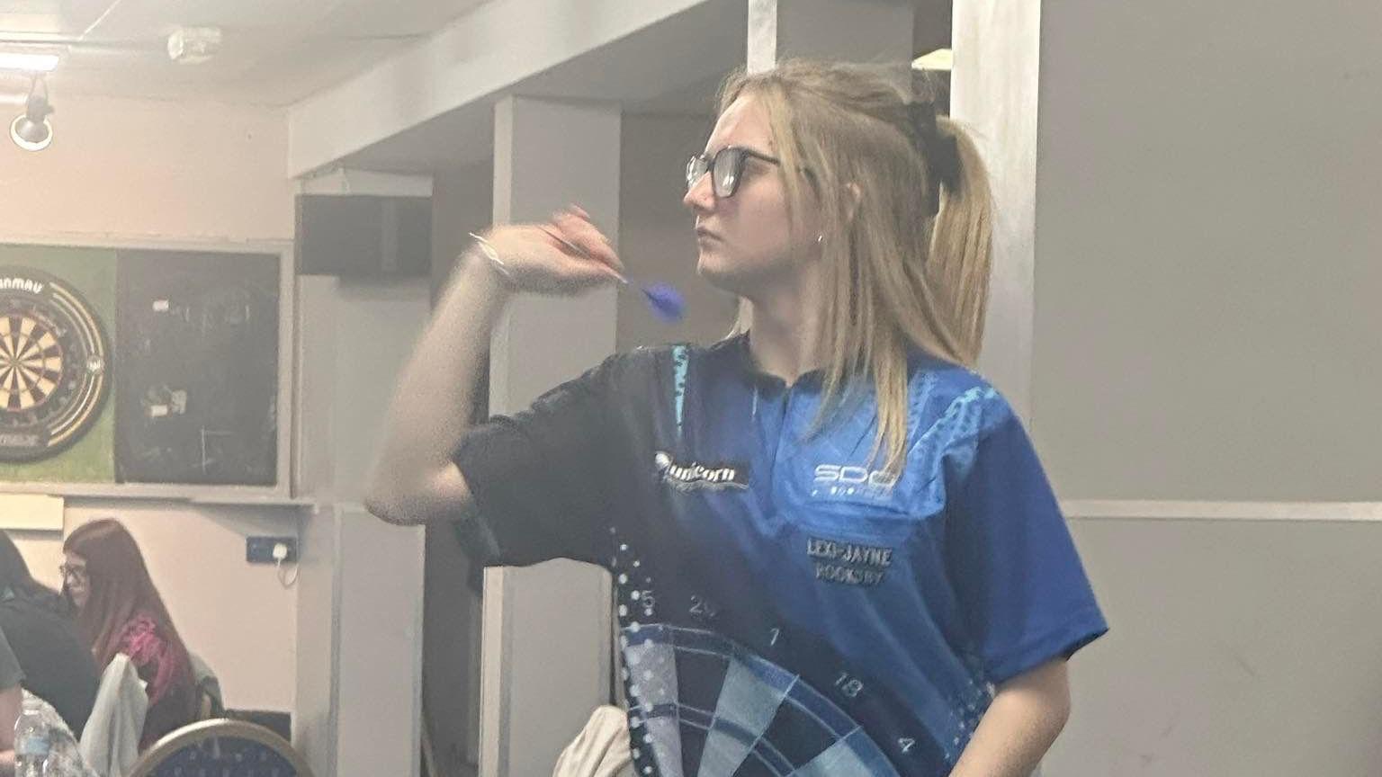 Girl wearing blue darts top prepares to throw dart at board