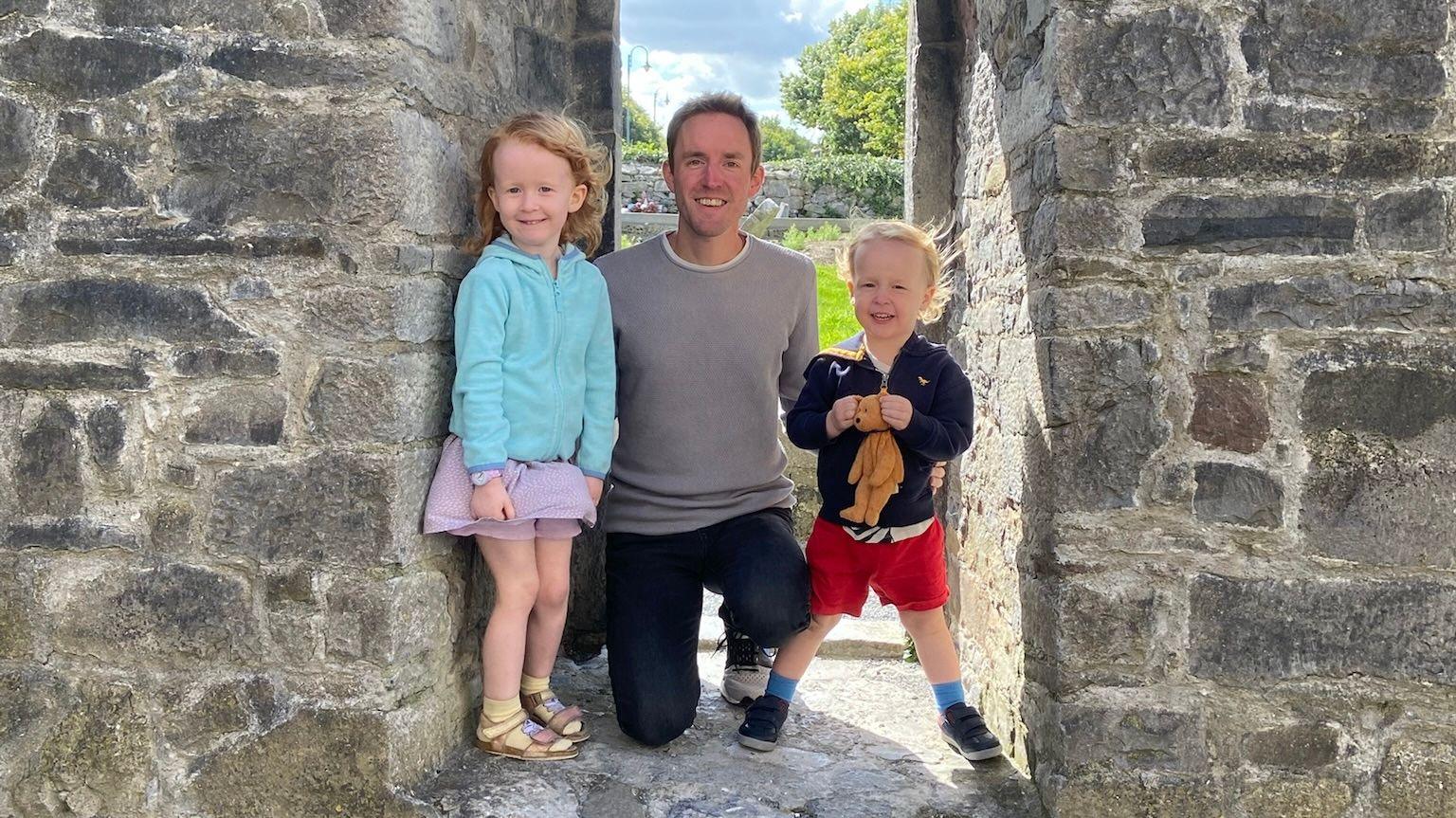 Mark pictured in the entrance to a grey castle with a young boy and girl who have strawberry blonde hair