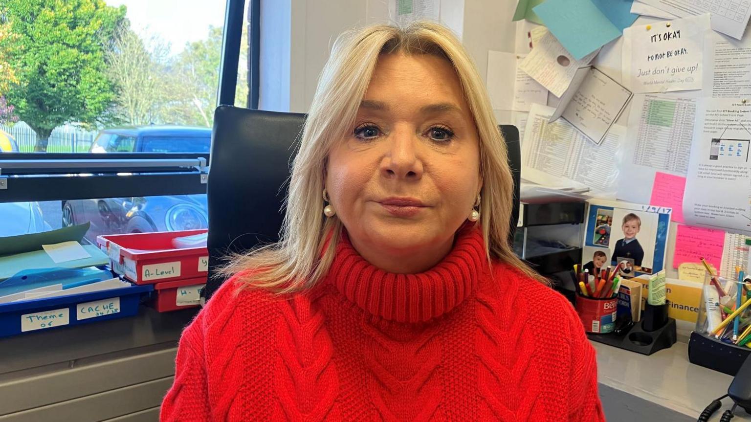 Ms Mackel is wearing a salmon coloured high necked jumper. She has shoulder length blonde hair. She's sitting in an office with files and pens behind her.