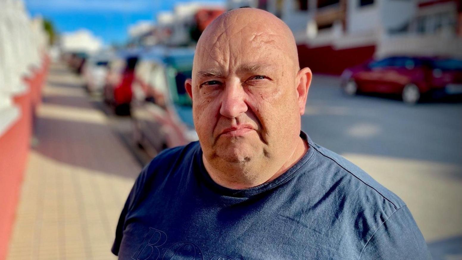 Billy George stands in the middle of the street looking at the camera. He is bald and is wearing a navy blue tshirt. The background behind him is blurred.