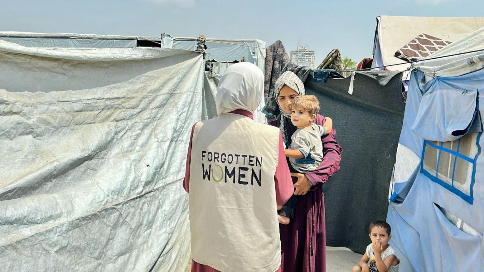 A woman, who is wearing a hijab and sleeveless jacket that reads, "Forgotten Women" on the back, is standing in front of a woman in a hijab holding a boy in her arms with another child crouching on the floor. There are makeshift tents on either side.