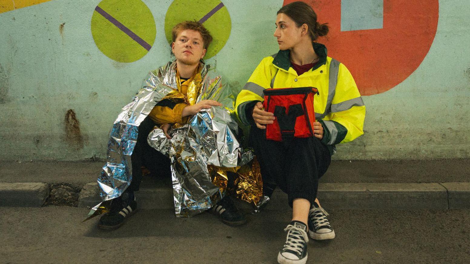 A man and woman sit on the ground against a wall painted with colourful geometric shapes. The man is wrapped in a metallic emergency blanket and wears black shoes. The woman is dressed in a high-visibility jacket and black trousers and is holding a red bag.