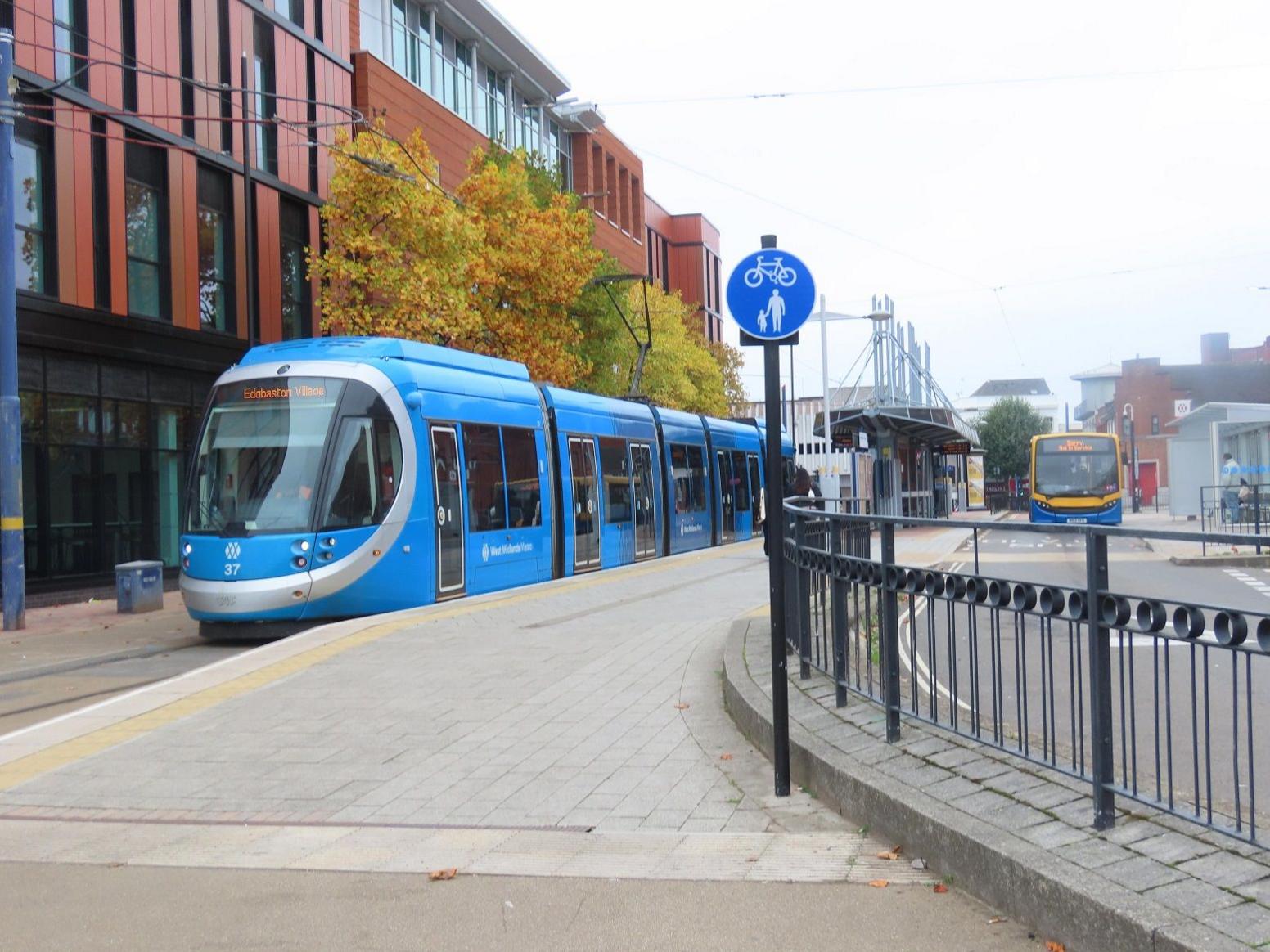 A blue and grey tram sits by a paved platform with a bus visible travelling alongside it in the distance.