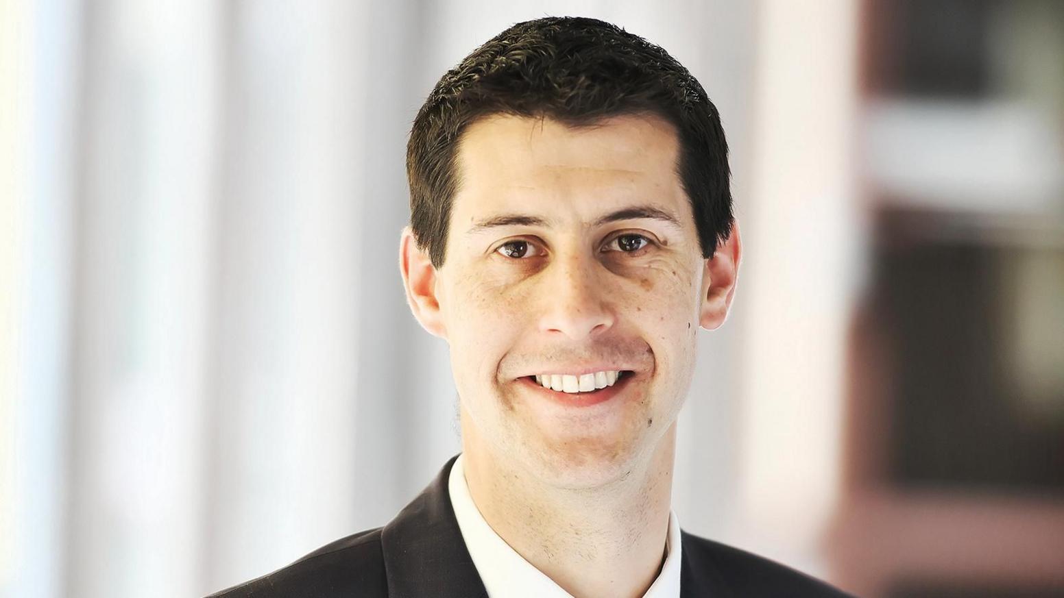 Head and shoulders shot of James Wilson, dressed in a suit and tie in a corridor at county hall in Norwich. He has short black hair and is smiling at the camera.