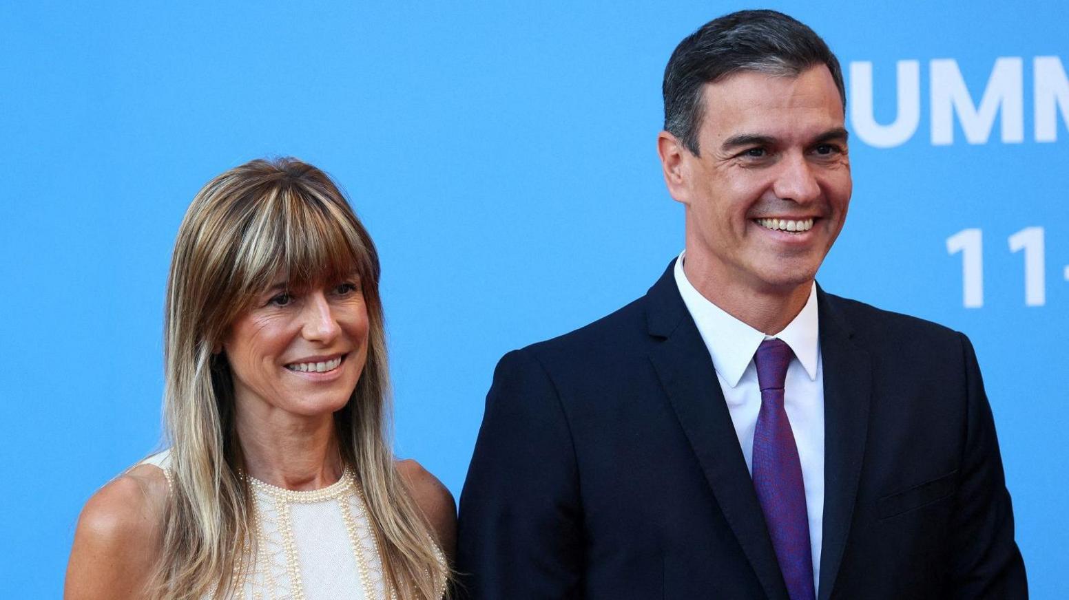 Sánchez and his wife Begoña Gómez are smiling with a blue background. Sánchez is wearing a suit and Gómez a white dress.