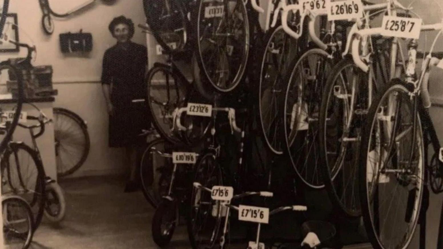 A woman is seen at the back of a shop, behind a display of bicycles in this black and white old photo. Prices on the bicycles are listed in pounds, shillings and pence.