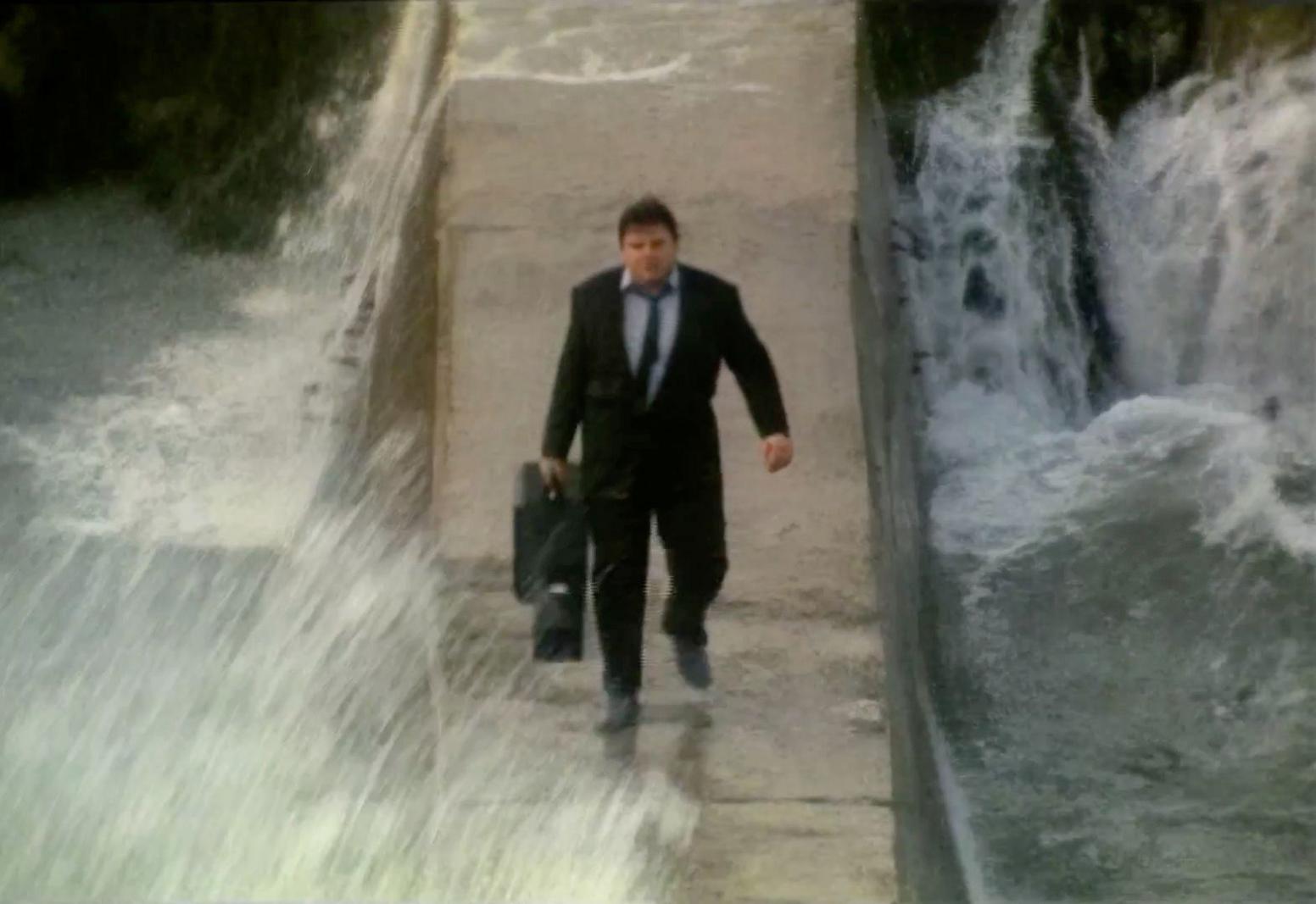 Actor Robbie Coltrane, wearing a suit and holding a guitar case, walking along the Hope Cove Harbour breakwater in strong seas and high waves in the film The Supergrass (1985).