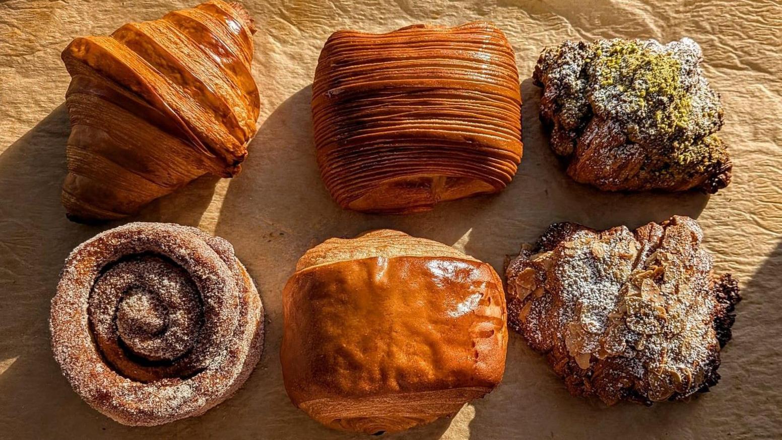 Six pastries are pictured in this photograph, including croissants, pain au chocolat and a cinnamon roll. They're displayed in two rows of three.
