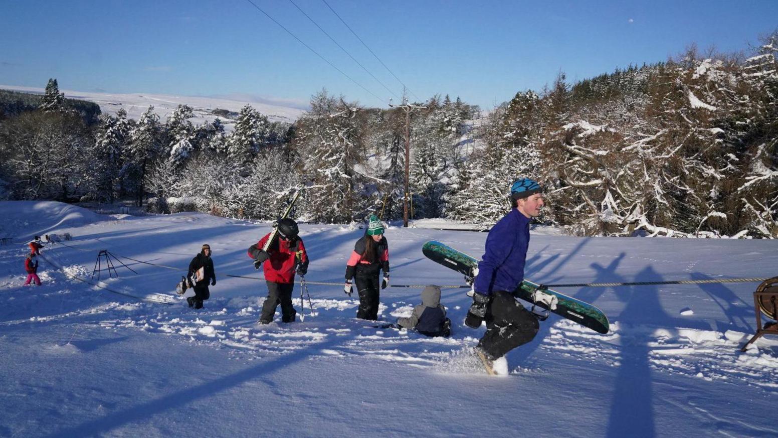 View of four people trooping up a snowy slope to the left of a rope-tow. They are carrying skis or snowboards and wearing appropriate clothing. Snow laden trees are behind them, with a hill in the background under a clear sky