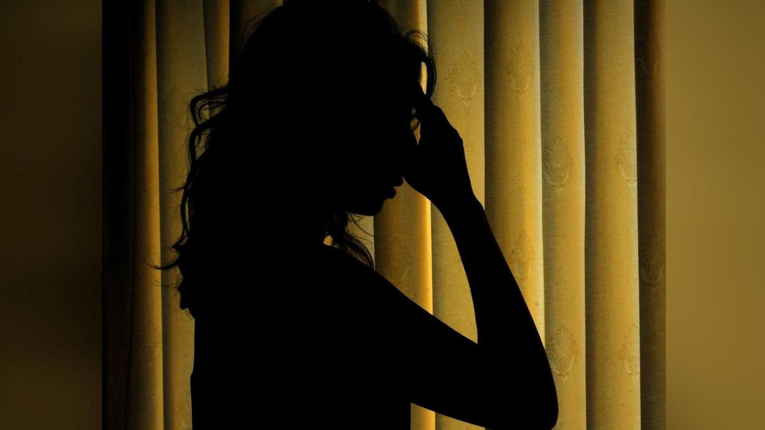 A stock image of the silhouette of a woman holding her hand to her face. The silhouette us set against the background of a mustard coloured curtain
