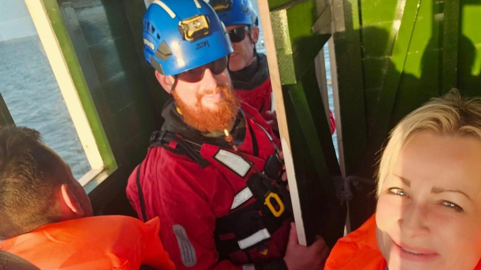 Two coastguards wearing blue helmets and life jackets coming through the door of the refuge. In one corner Dita Sanchez is smiling at the camera.