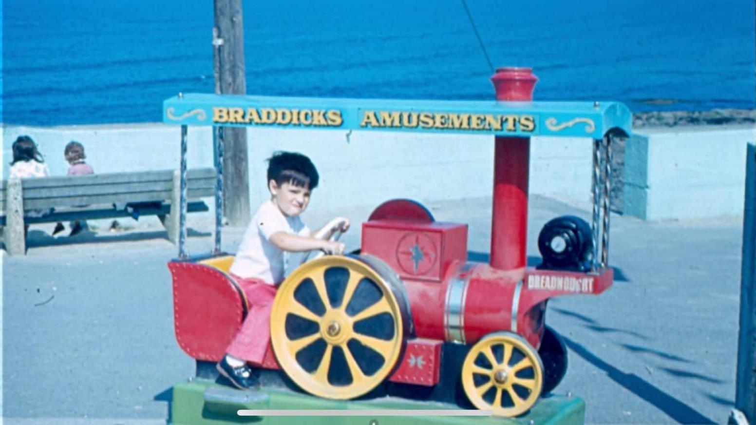 Steve Dance as a young boy is sitting on the seat of a toy steam engine. He has short dark hair and is wearing flared pink trousers and a white T-shirt with blue sandals. He has a scrunched up face and is holding the steering wheel of the ride. Across the the top of the ride is lettering saying: BRADDICKS AMUSEMENTS. It sits on the pavement and there is also a bench with a woman and a child sitting on it. The beach and sea are visible behind the pavement.