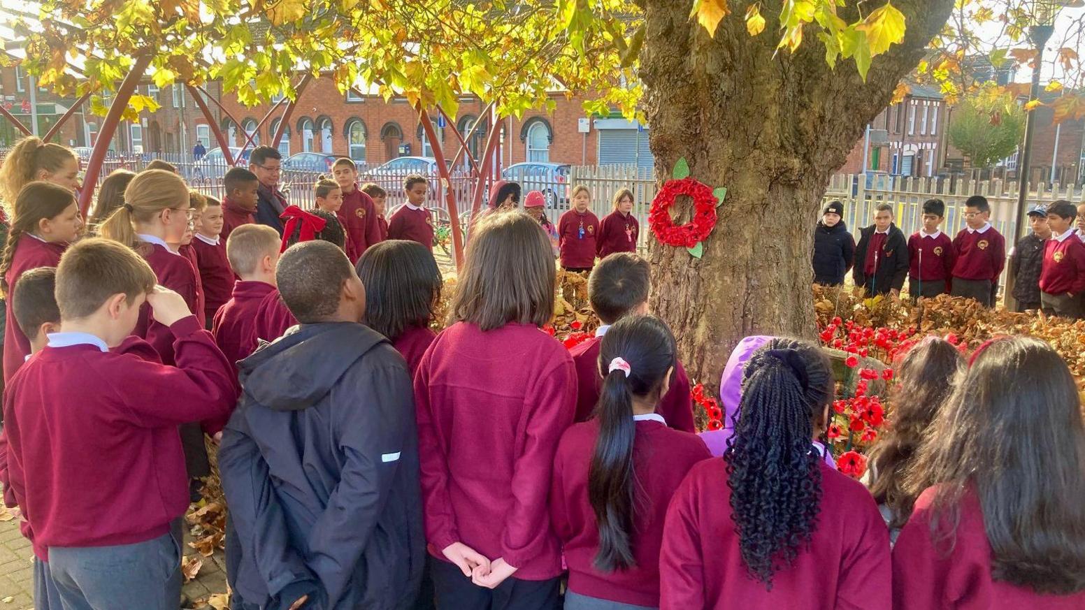 A group of about 30 primary school girls and boys stand in a circle around a tree which has a poppy wreath on the trunk and plastic poppies around the base. You can see a few green / yellow leaves in the top of the photo. The children wear grey school trousers and a maroon jumper. They are all looking inwardds towards the tree.