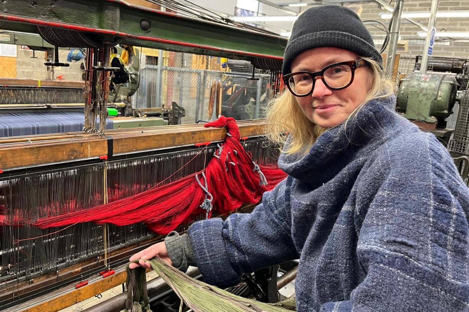 Clare is working at a loom at her business in Evanton. She is wearing a thick woolly coat and a hat.