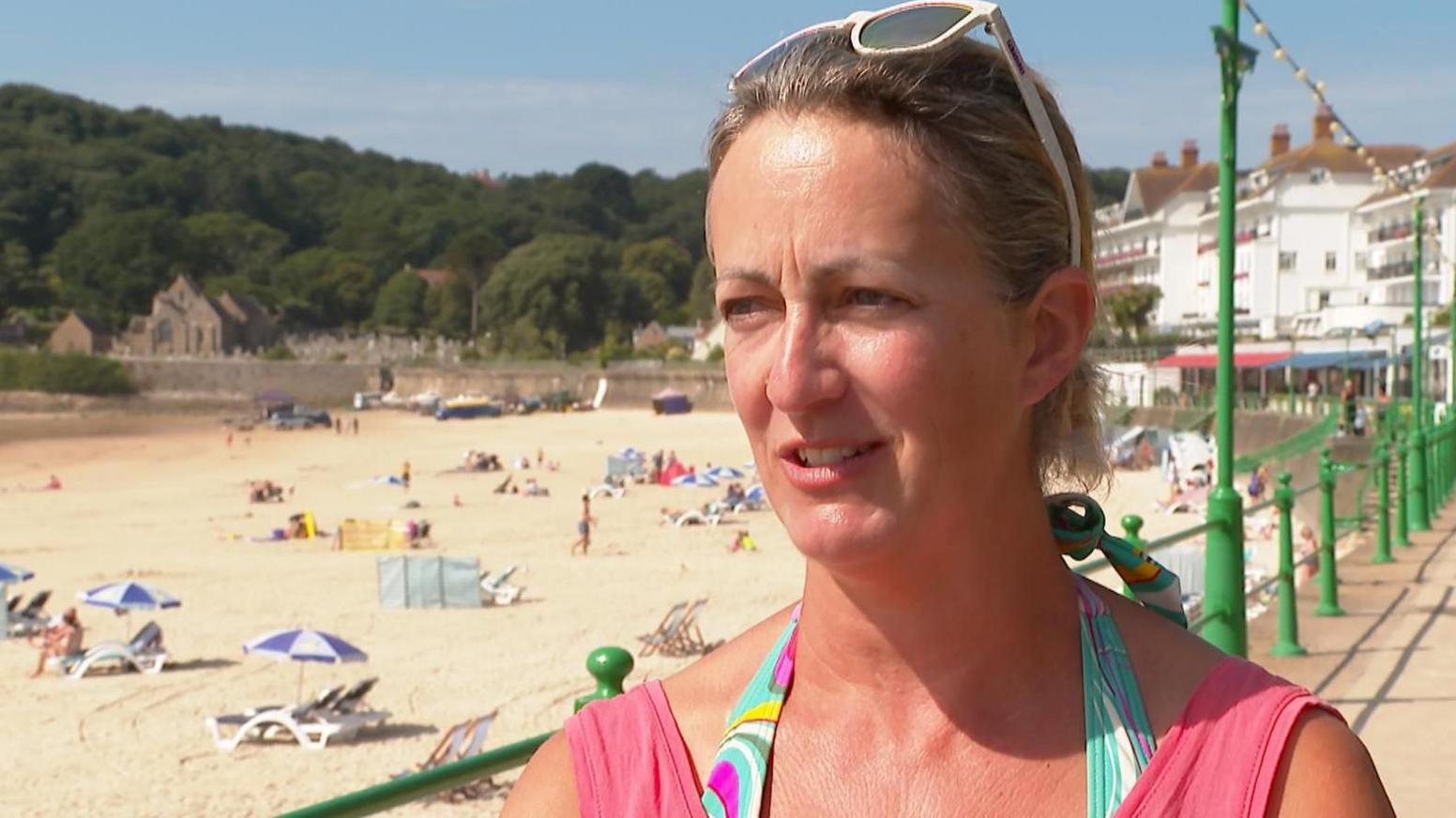 Holidaymaker Ginnie wearing a pink T-shirt. She has sunglasses on her head. She is standing on a promenade with a beach in the background filled with people sunbathing.
