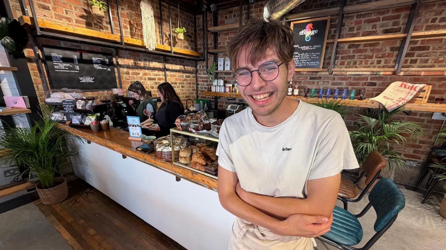 Cafe worker Ryan Stewart who is wearing thin-rimmed black glasses and a white Lee Cooper T-shirt is smiling with his arms crossed. He is standing in front of the till area where a number of cakes are in display cabinets. In the background are two of his colleagues, one is a woman with long black hair and the other has shorter hair and is wearing a green jumper.