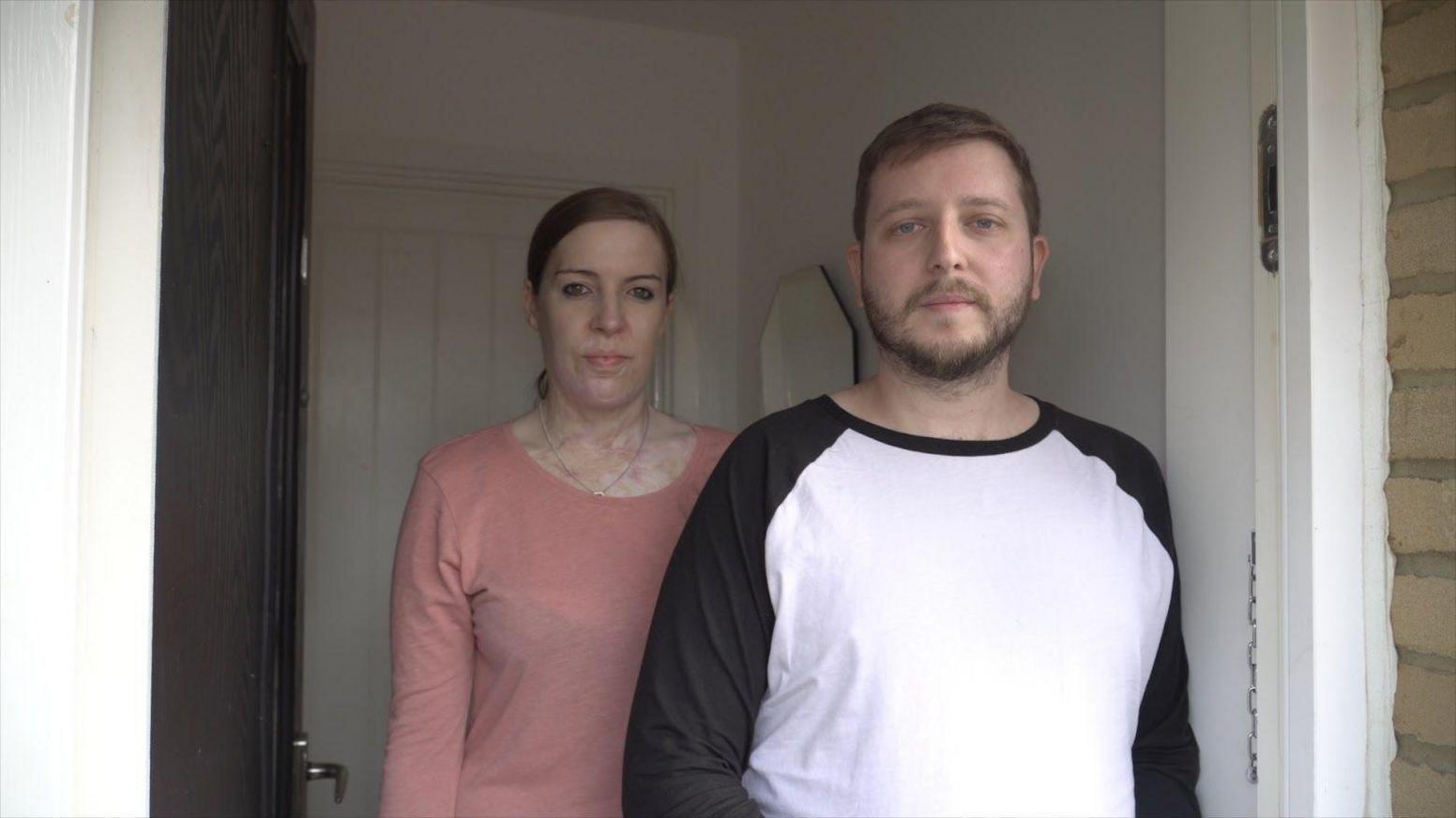 A man wearing a black and white t-shirt and a woman wearing a pink t-shirt stood in a doorway. They look cross.
