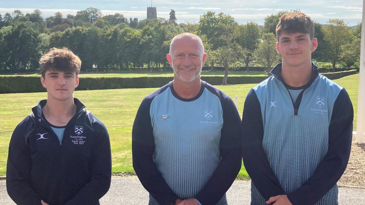 Felix Lagerberg, Will Cotterill and Dan Graves smile and look directly at the camera as they pose in a line. They all are holding their hands together in front of them and wearing Framlingham College sports kit. There is a field behind them, with trees and the top of the church. 