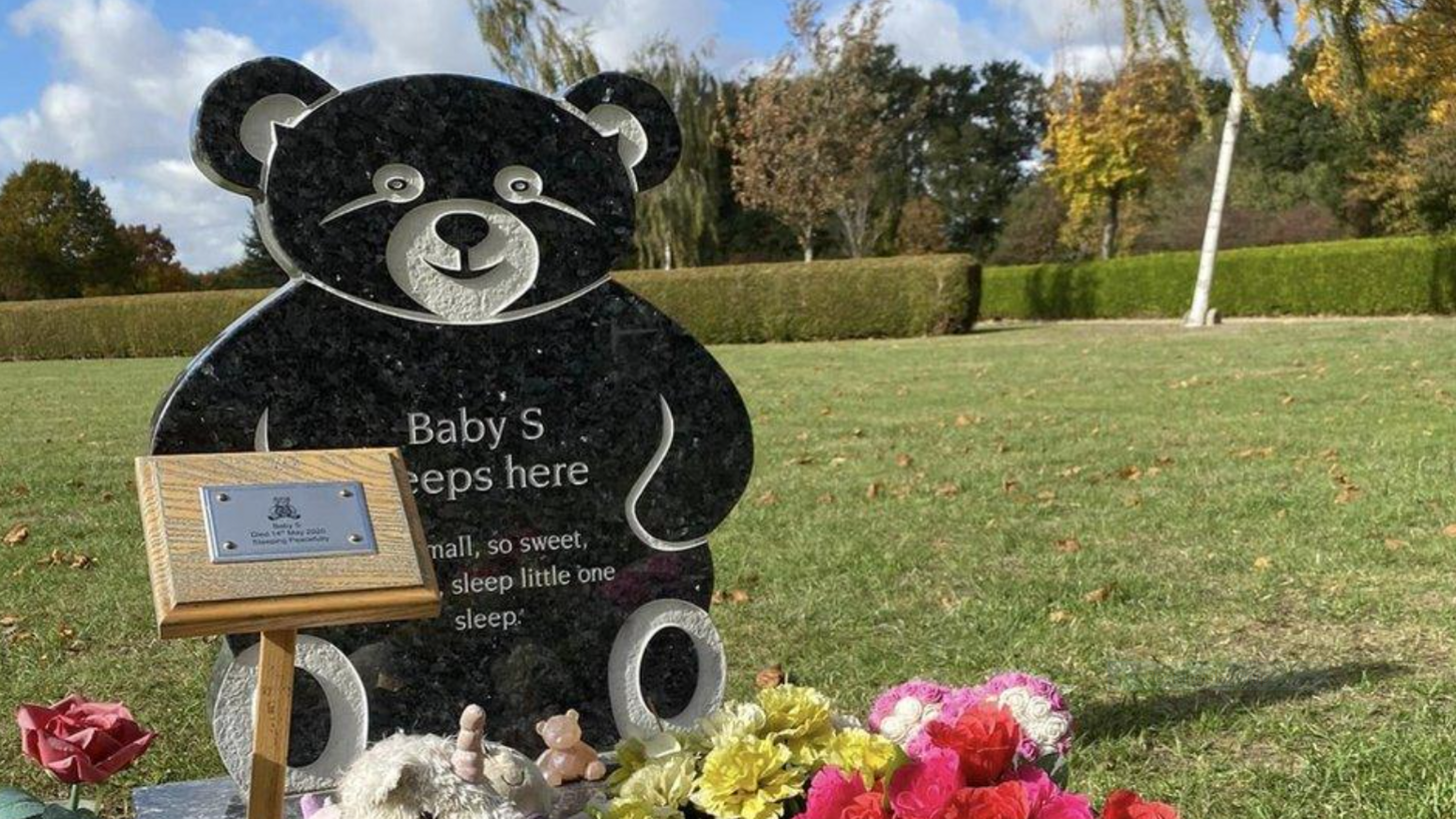 A large bear-shaped headstone with the Baby S written on it. It is positioned in a field and in front of some flowers and a wooden and mental plaque
