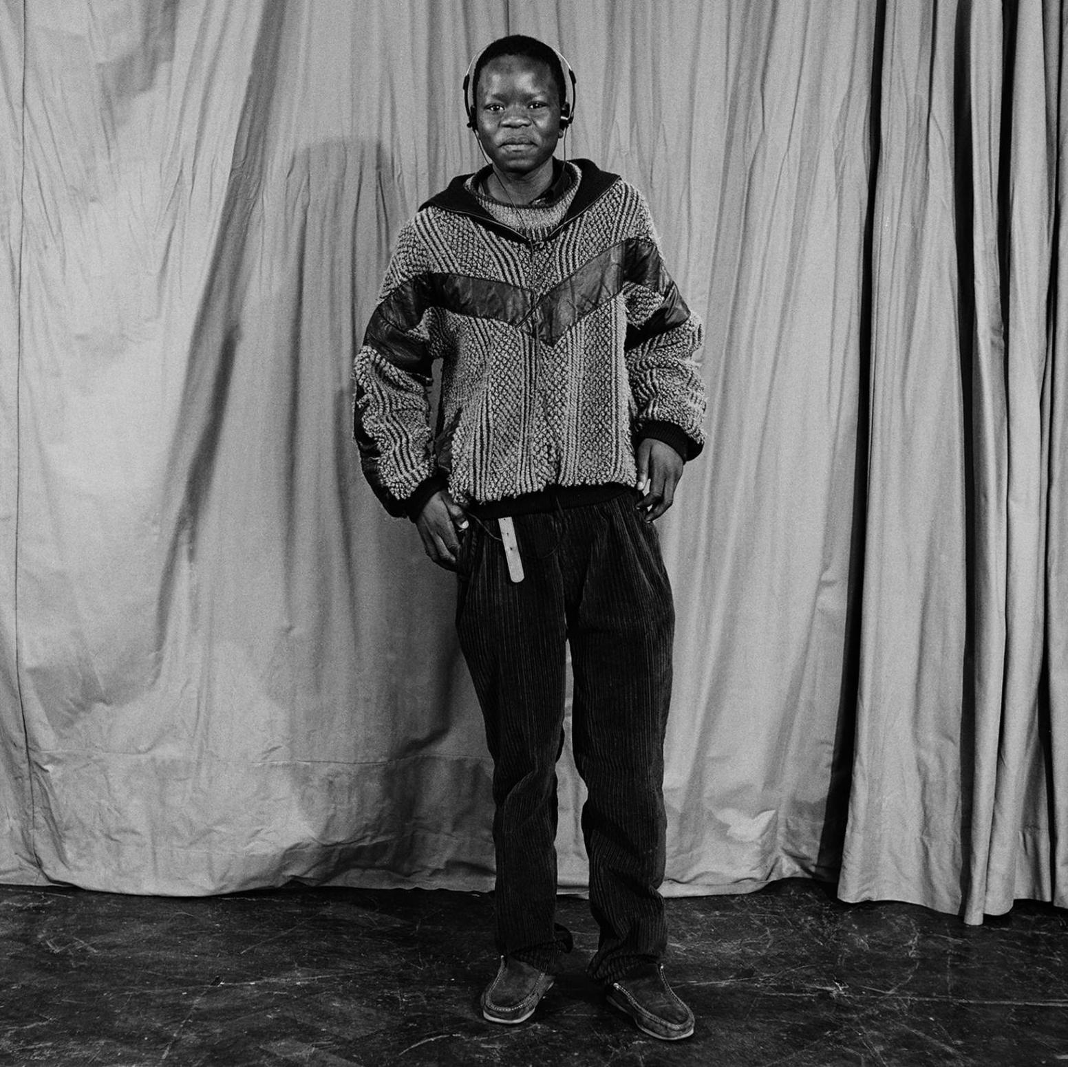 Boys from Tulse Hill School, 1990. A young men stands in front of a curtain. He wears headphones, a patterned jumper ans dark trousers. Black and white photo.