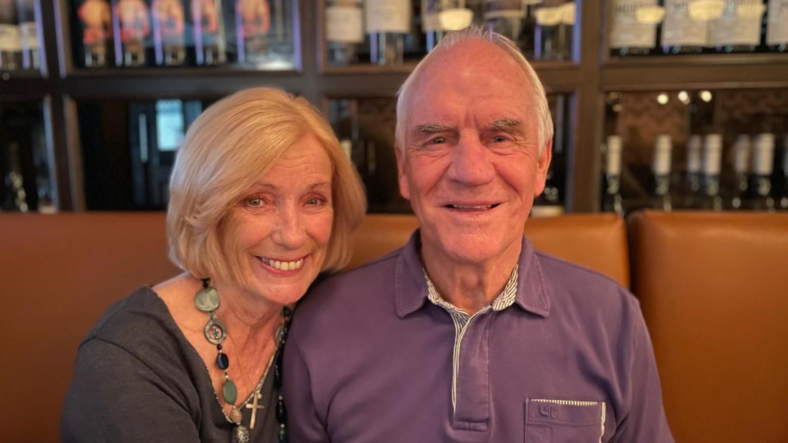 Penelope and Dave Watson, sitting side by side on a sofa in a bar, smiling at the camera