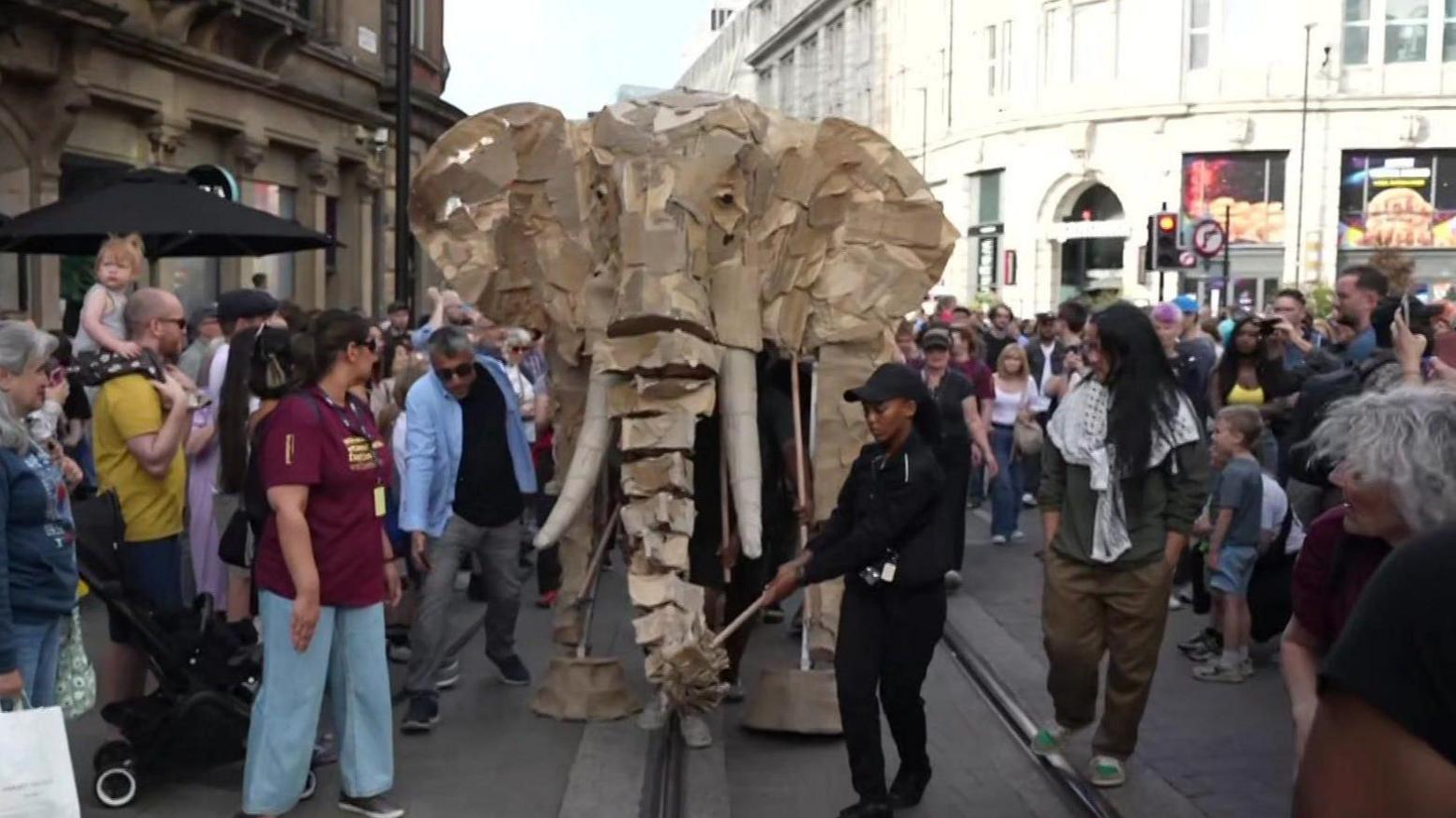 A wooden elephant puppet moves through Manchester city centre with crowds around