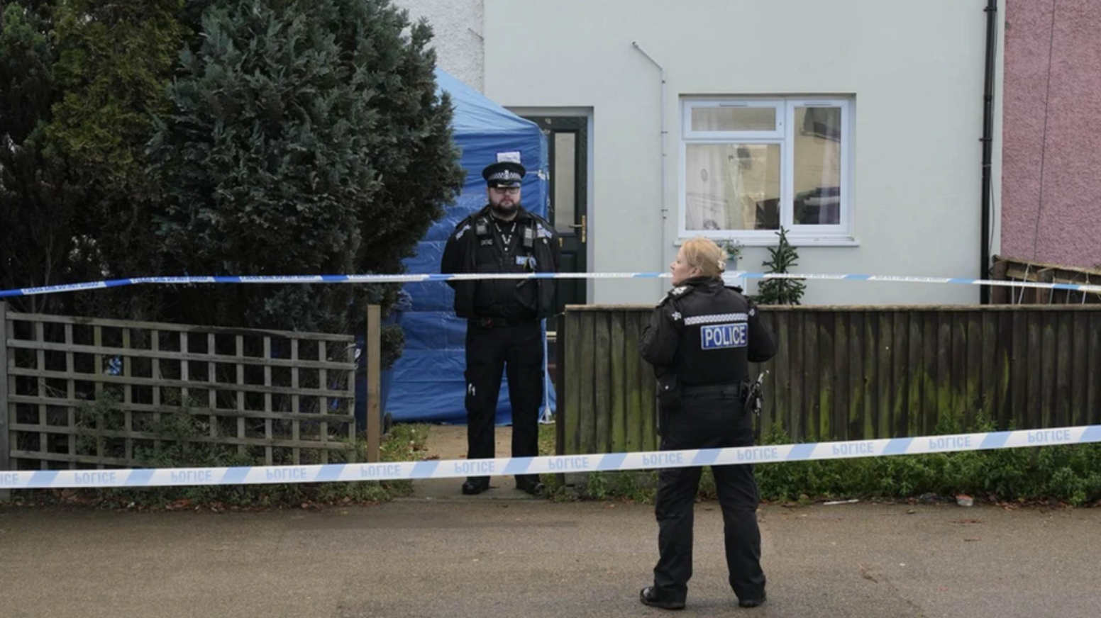Two police officers standing outside a small house. A blue tent is in the front garden and police tape is around the scene.