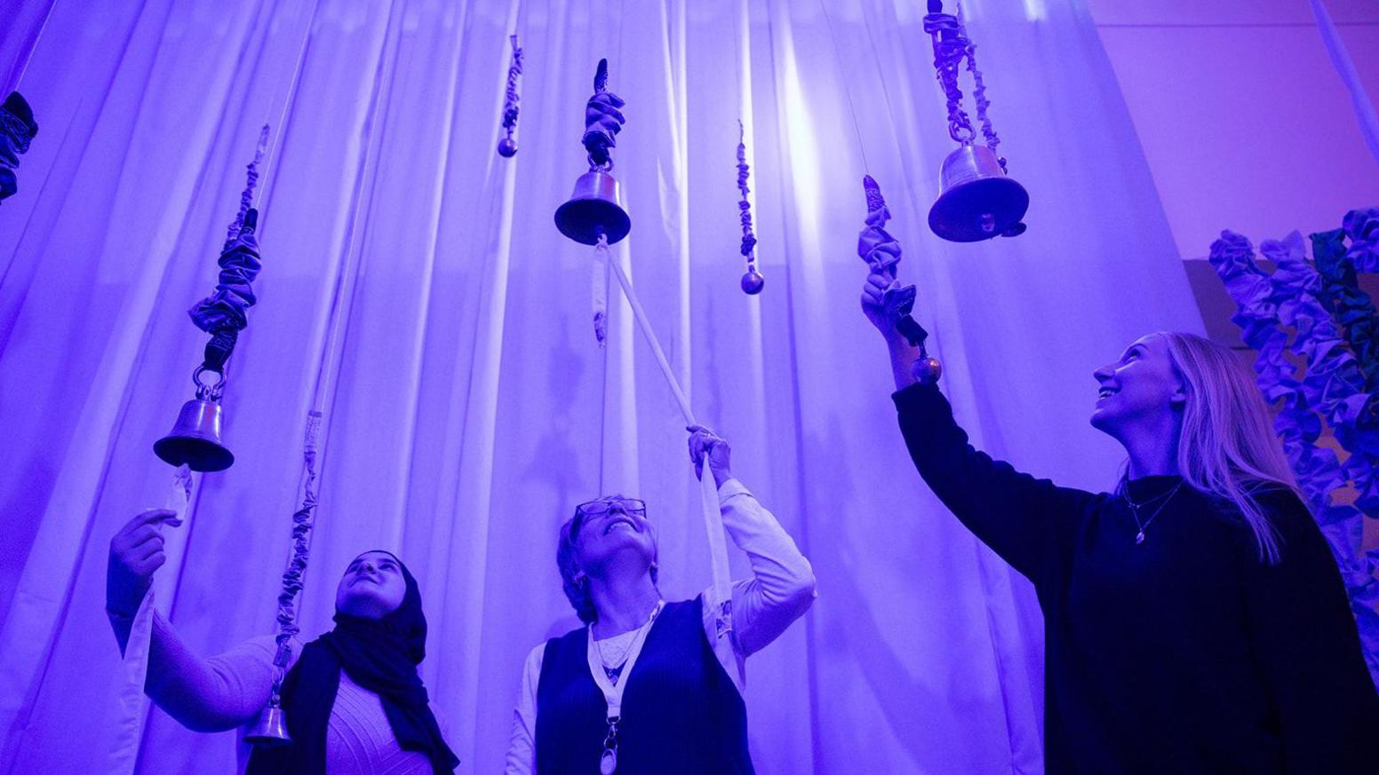 Bells hang from the ceiling as three women have a go at ringing them.