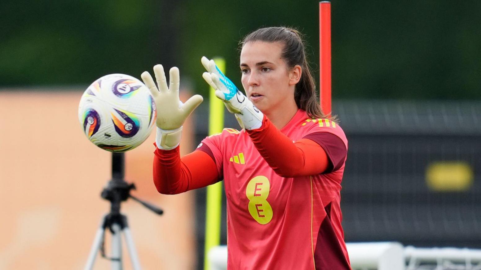 Olive Clark, with brown hair in a ponytail, has her arms out in front of her. She is about to catch a ball which is travelling towards her. She is wearing a red football shirt with EE in yellow letters. She is wearing white goalkeeper gloves.