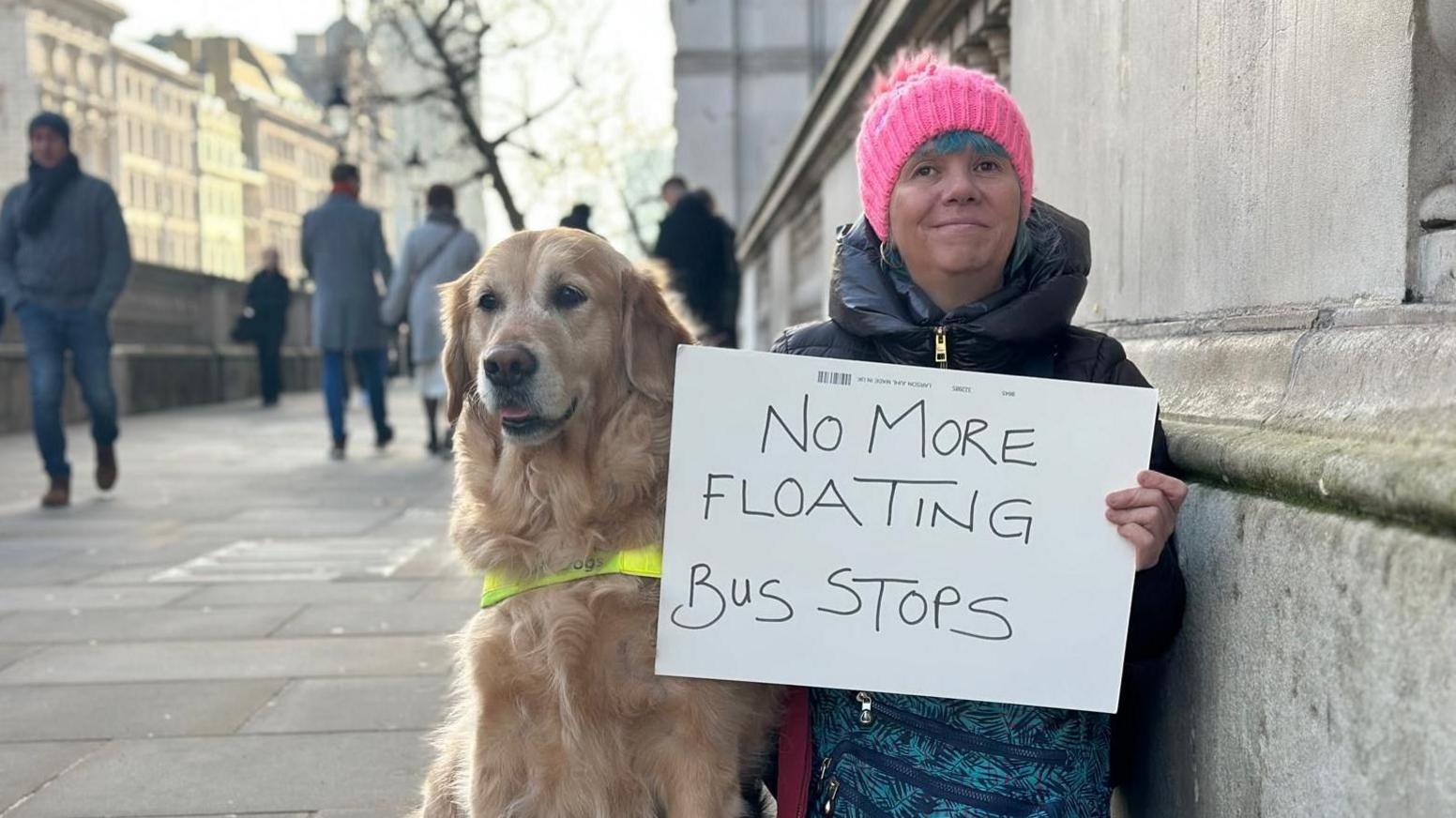 A woman wearing a pink hat, crouched against a stone wall, holding a white sign that says "no more floating bus stops". Sat next to her is a golden retriever guide dog. People can be seen walking the pavement next to them.  