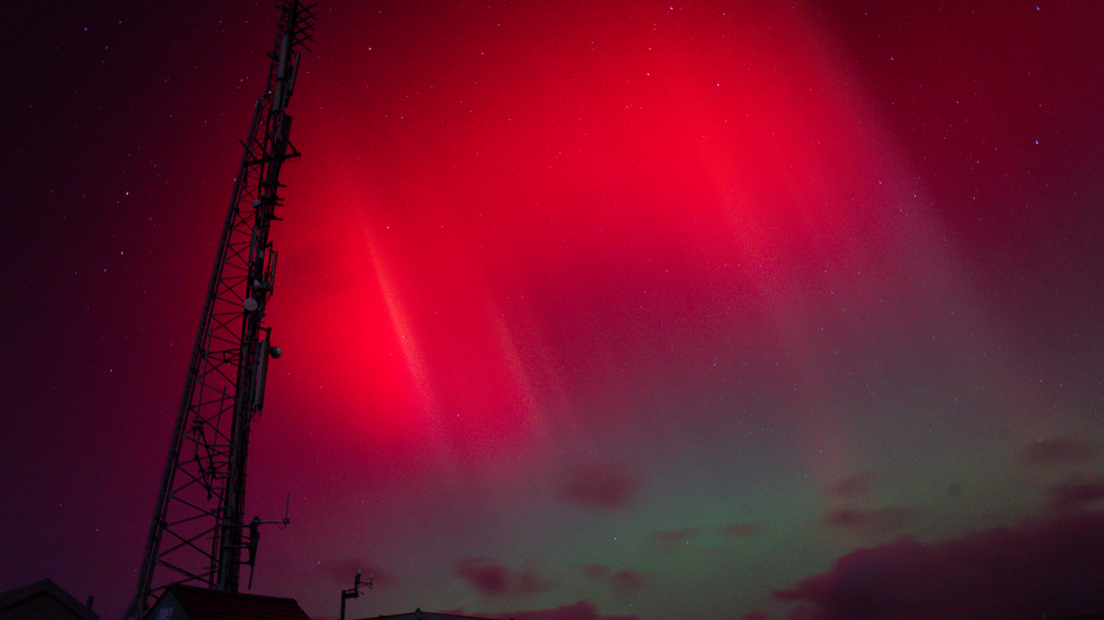 A telecoms tower stands in the foreground, backlit by bright red shafts in the night sky. Dappled clouds float in deep blue and green sky to the right,