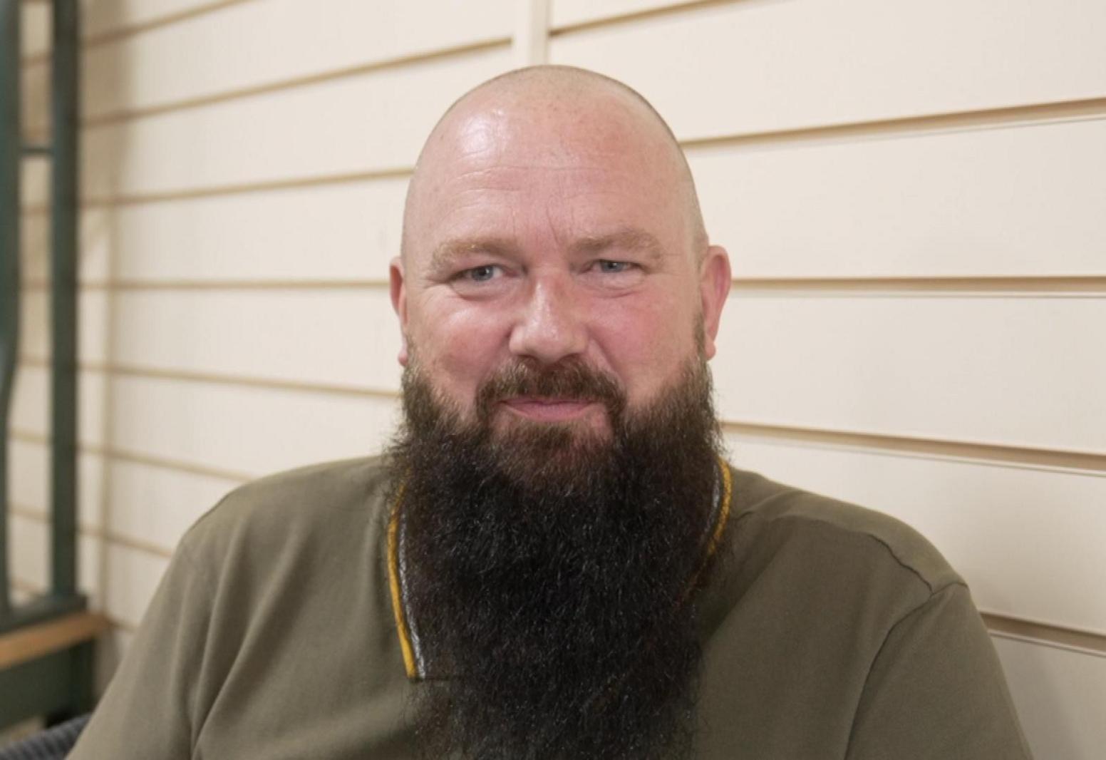 A man with a long black beard and a olive polo shirt smiles at the camera 