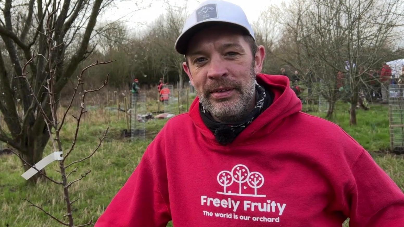 James wears a Freely Fruity hoodie and grey cap, and stands next to a newly-planted tree.