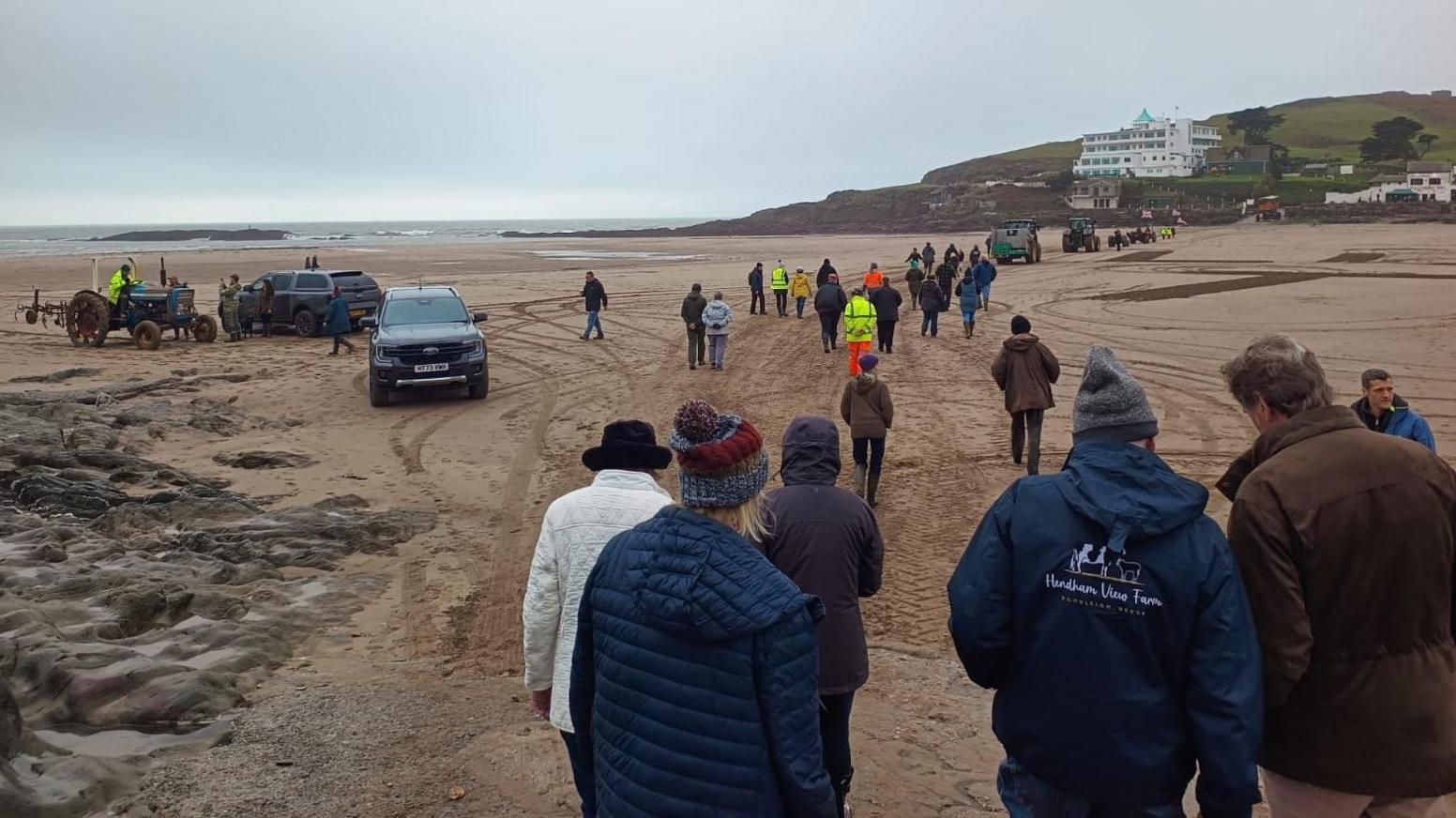 Dozens of farmers gather on Bigbury Beach in Devon. The group has its back to the camera and is walking off into the distance across the beach. The weather is grey and there are various vehicles and tractors parked on the sand. 