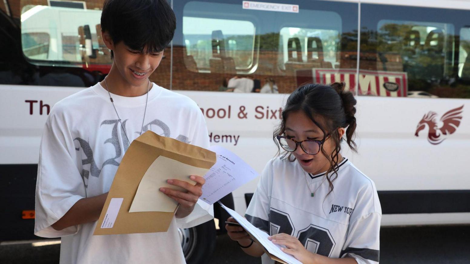 Two students stand in front of a school minibus opening their results. The student on the left, a male, has short black hair and wears a baggy white short sleeve t shirt with silver graffiti writing on the front. He is holding a brown envelope and smiling as he looks down at the paper. To the right of the image is a young woman who has her mouth open in happy surprise. She has black hair tied back in a messy bun, with two curly strands framing her face and wearing thick framed black glasses and a white and black American football t shirt.