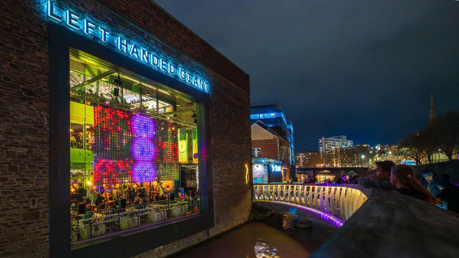 A building is lit up by a light installation in the window. The building has a sign that says Left Handed Giant.