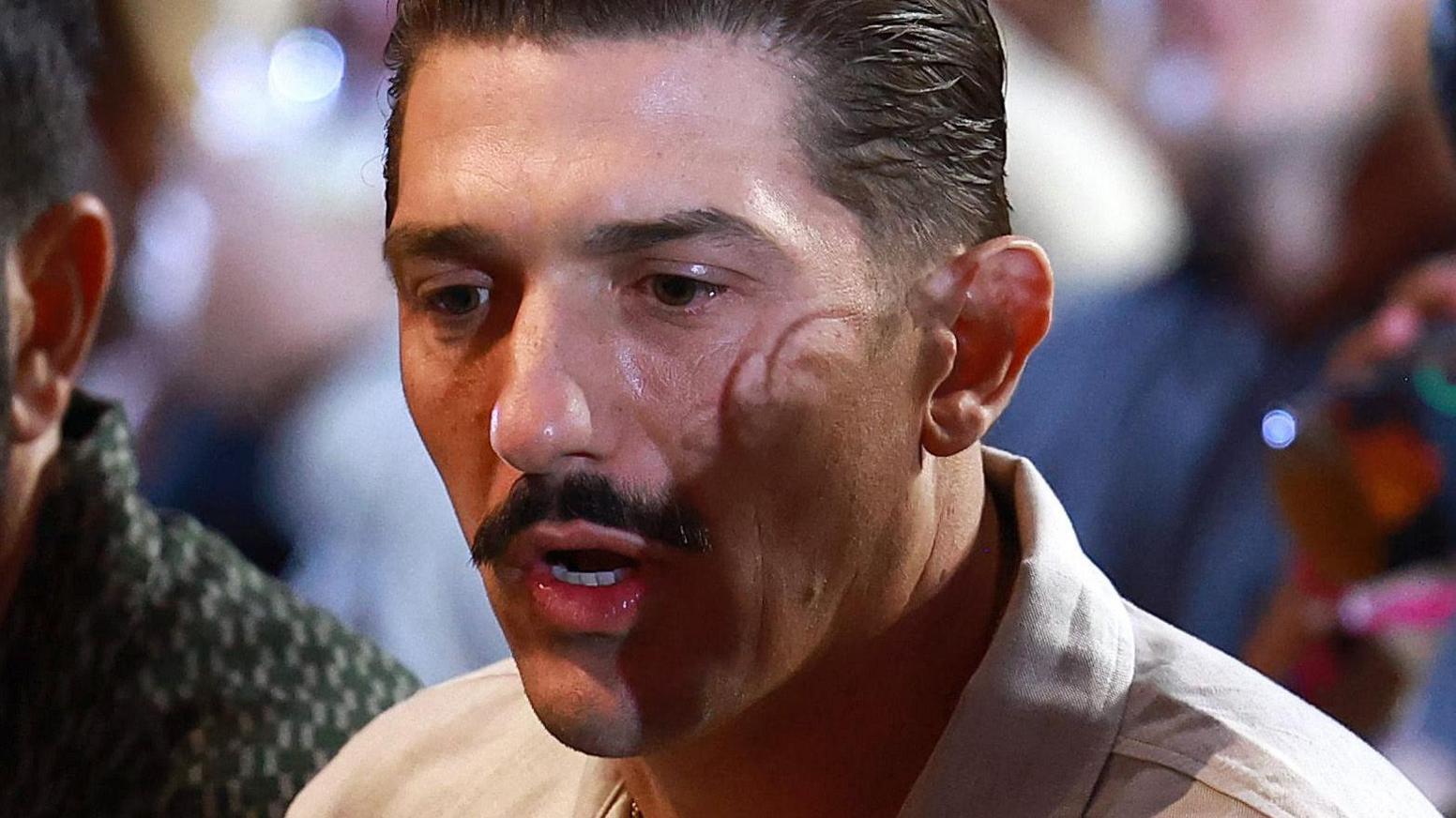 Andrew Schulz pictured at an event wearing a white shirt. He has slicked back dark hair and a moustache.  