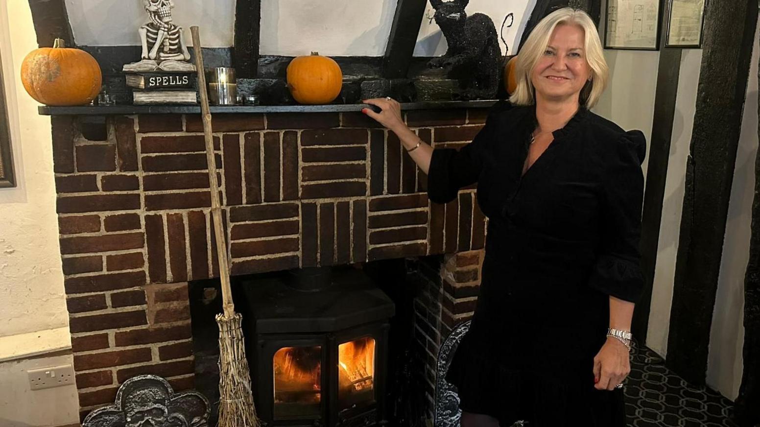 A woman with shoulder length blonde hair is smiling into the camera as she stands in front of a large red and white brick fireplace. She is wearing a black dress with elbow length sleeves and rests her right arm on top of the mantlepiece on which sit Halloween decorations including two orange pumpkins, and pile of books with one titled 'Spells' and a witches' style broom leans against the hearth.