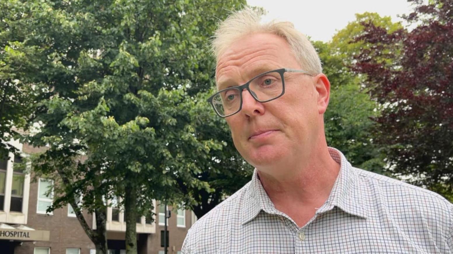 Patrick Armstrong - a man with light blond/white hair with dark rectangle glasses, wearing a white chequered collared shirt. Behind him is the hospital building to the left and trees in front and to his right.