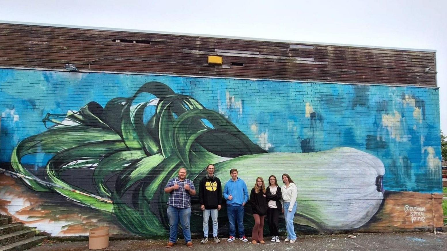 Six people standing in front of the mural, which is of a giant leek.