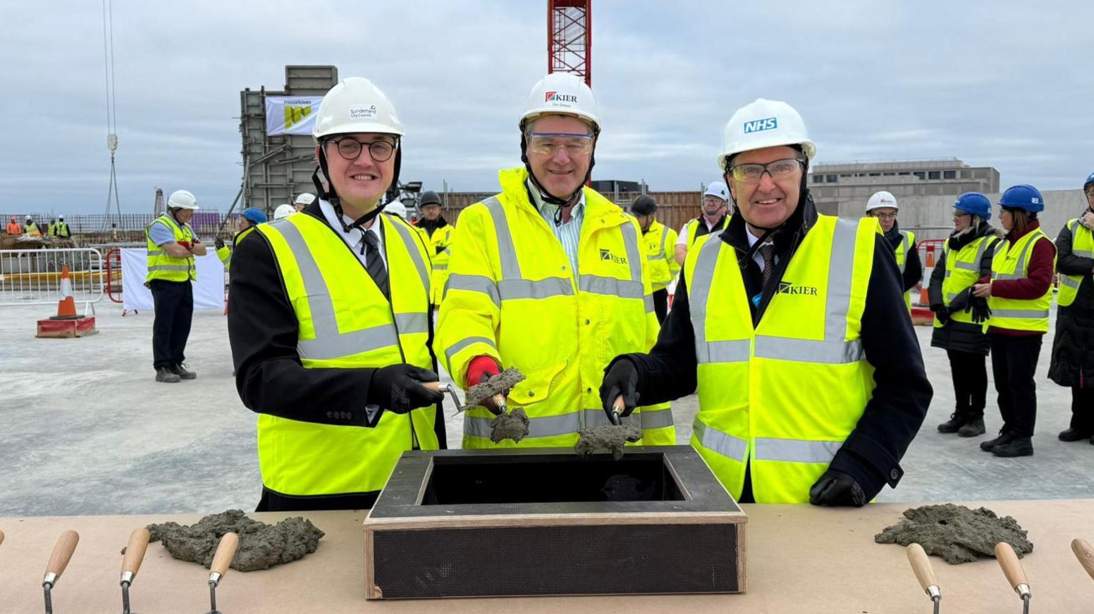 Three men wearing white hard hats and yellow high visibility jackets hold up a trowel containing cement before placing it into a black box.