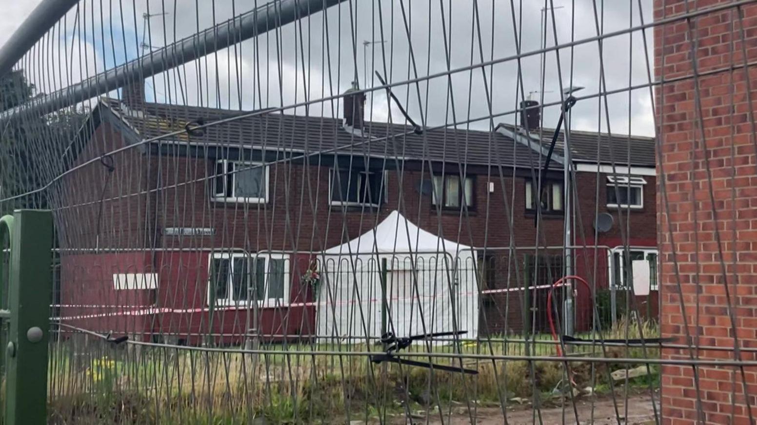 A brown block of flats can be seen through a wire fence. A white police tent, which is surrounded by police tape, has been set up next to the building.