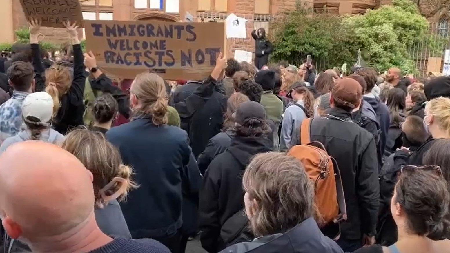 A crowd of protestors gather outside a former church building, one holding a sign saying "Immigrants welcome, racists not"