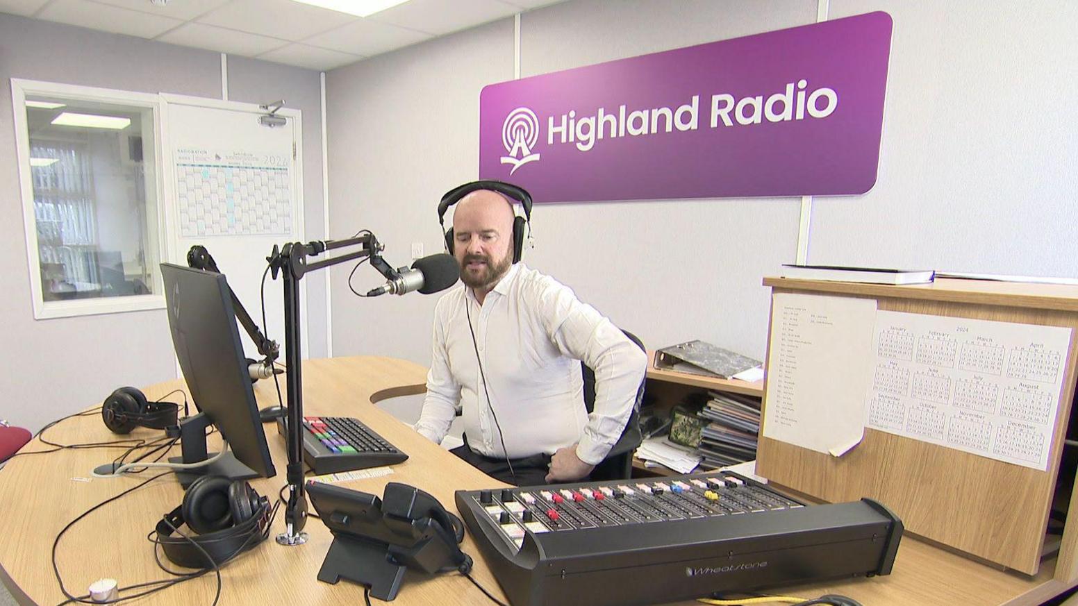 Greg Hughes - a man wearing a white collared shirt is sat at a large wooden desk. He is wearing black, overhead headphones and is sat in front of a computer screen, a black microphone and a mixing desk. Behind him is a large purple sign with 'Highland Radio' written in white font.