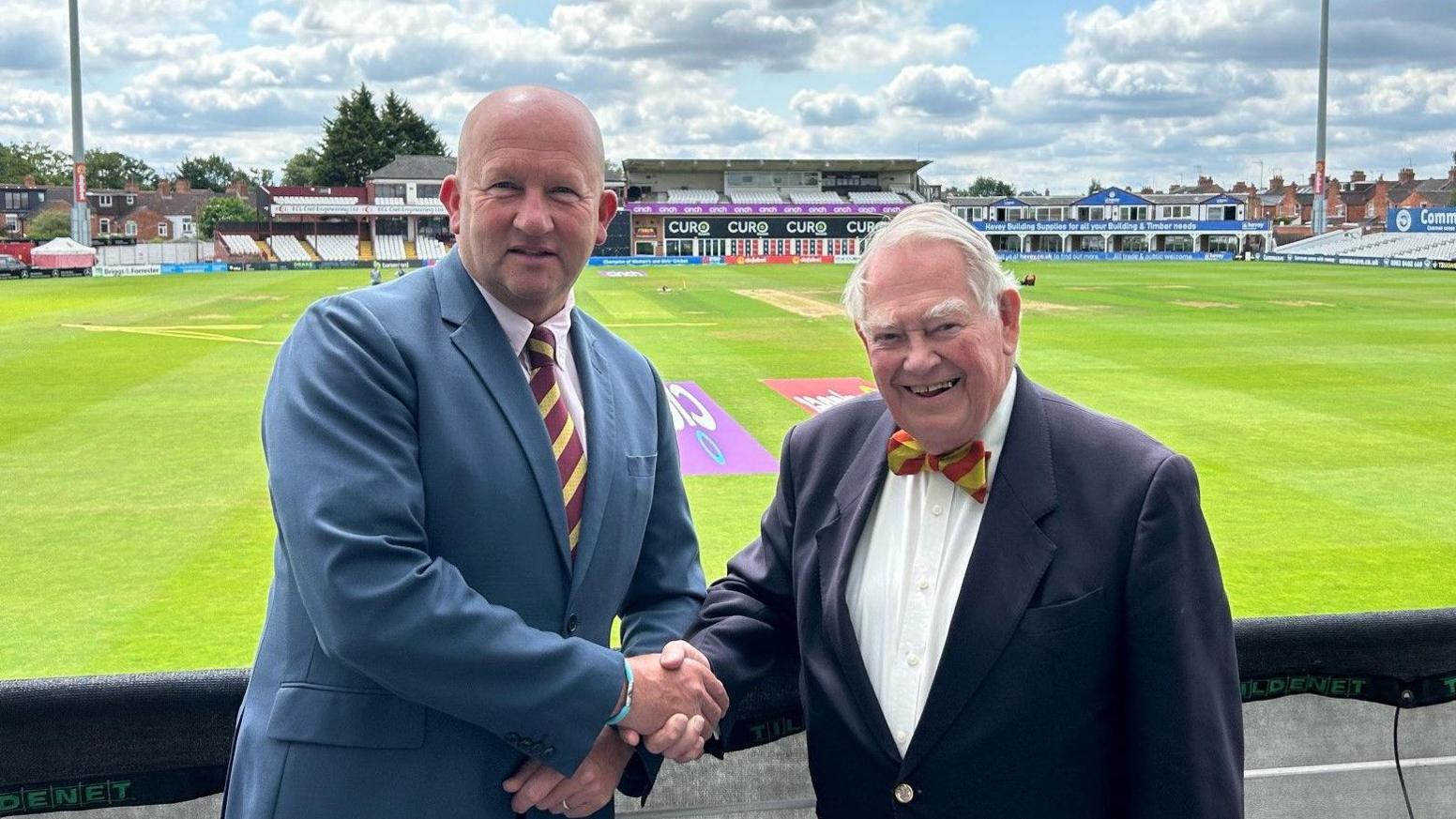 Gavin Warren and Lord Naseby shaking hands with a cricket pitch in the background