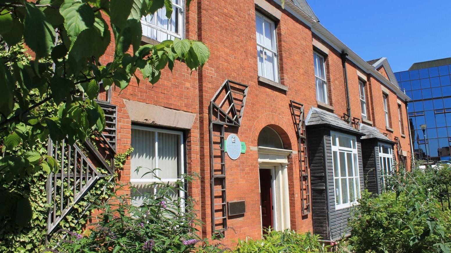 The outside of the Pankhurst museum, which is made up of two red brick Georgian semi-detached villas with green foliage outside. It is draped in sunlight.