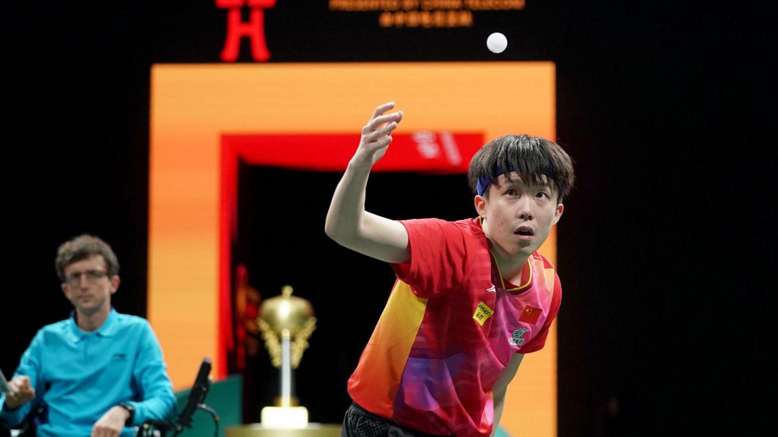 Mr Foote in a blue shift in the background sat in an umpire chair. Wang Chuqin in in the middle of a match in the foreground wearing a red t shirt. 