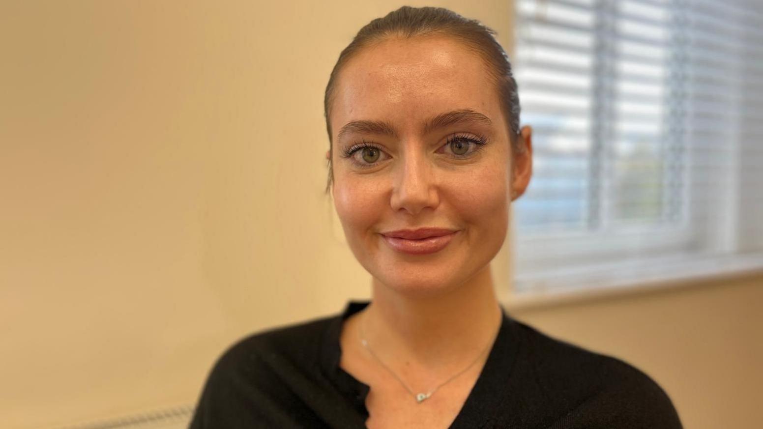 Lydia is wearing a black top and is smiling at the camera. Her dark hair is pulled back into a bun and she is sat in a room with cream walls and a window behind her. It is a head and shoulders shot. She is in focus while the background is slightly blurred.