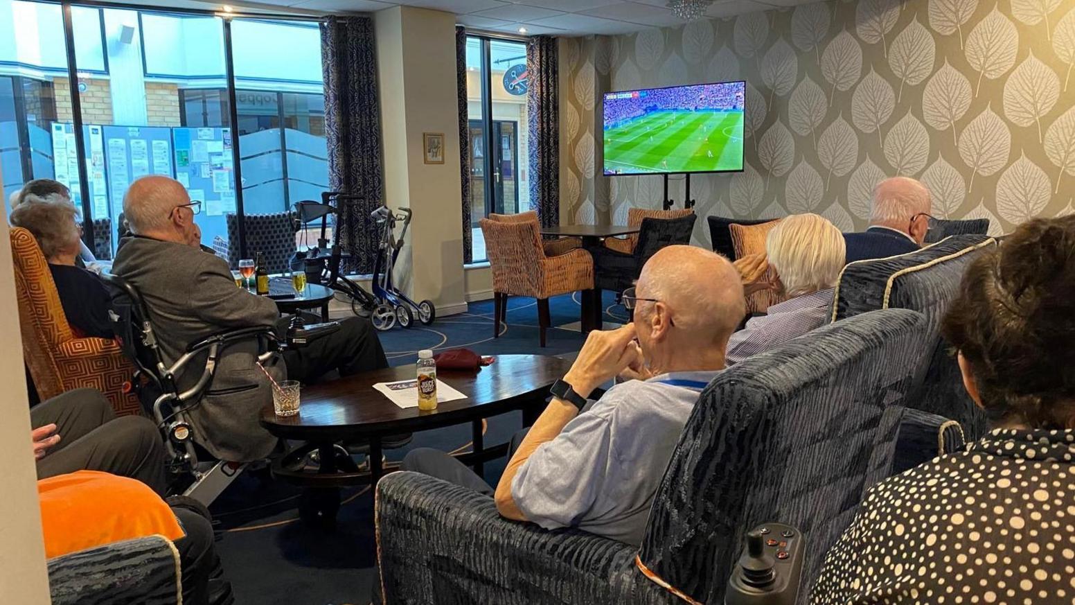 Residents watching the game in Lichfield. Several men and a woman are sat with their backs to us in several arms chairs around a wooden table with a flat screen TV showing the match on the wall