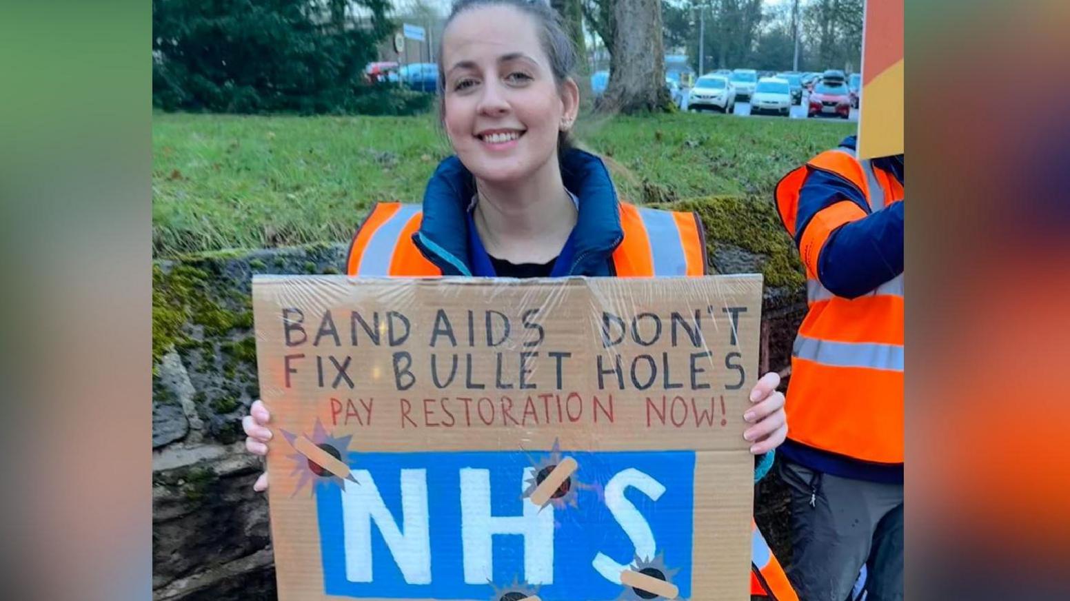 A woman with her hair pulled back in a pony tail and wearing an orange hi-vis tabard holds up a placard which reads "band aids don't fix bullet holes. Pay restoration now. NHS".